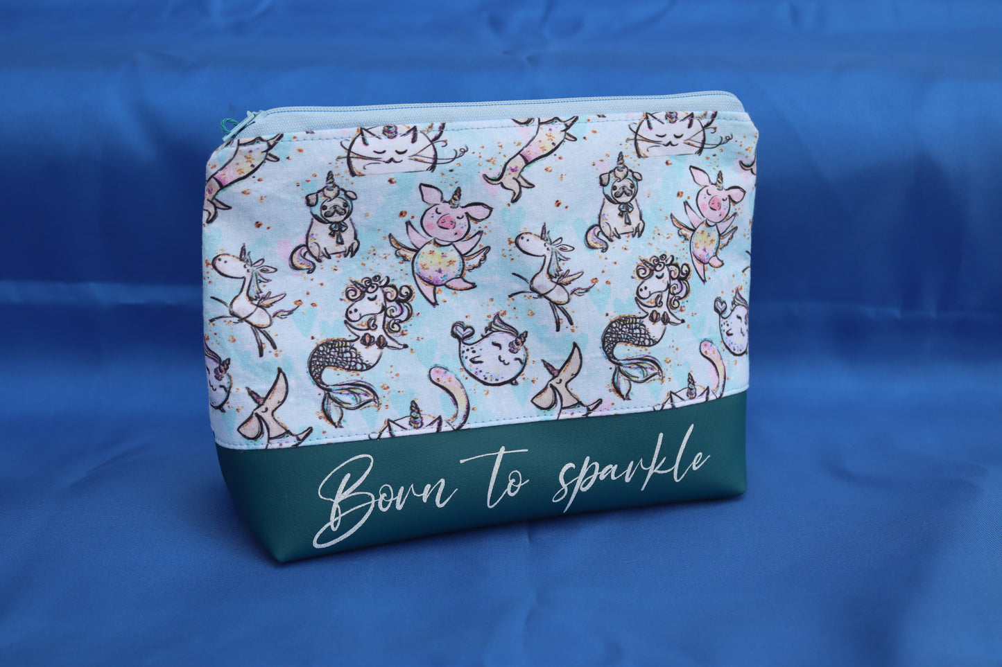 Handgemachte Kosmetiktasche mit hellblauem Stoff und niedlichen Fantasietieren wie Einhörnern, Meerjungfrauen, Mops-Einhörnern und Schweinchen mit Flügeln, darunter petrolgrünes Kunstleder mit der Aufschrift „Born to sparkle“, fotografiert vor blauem Hintergrund.