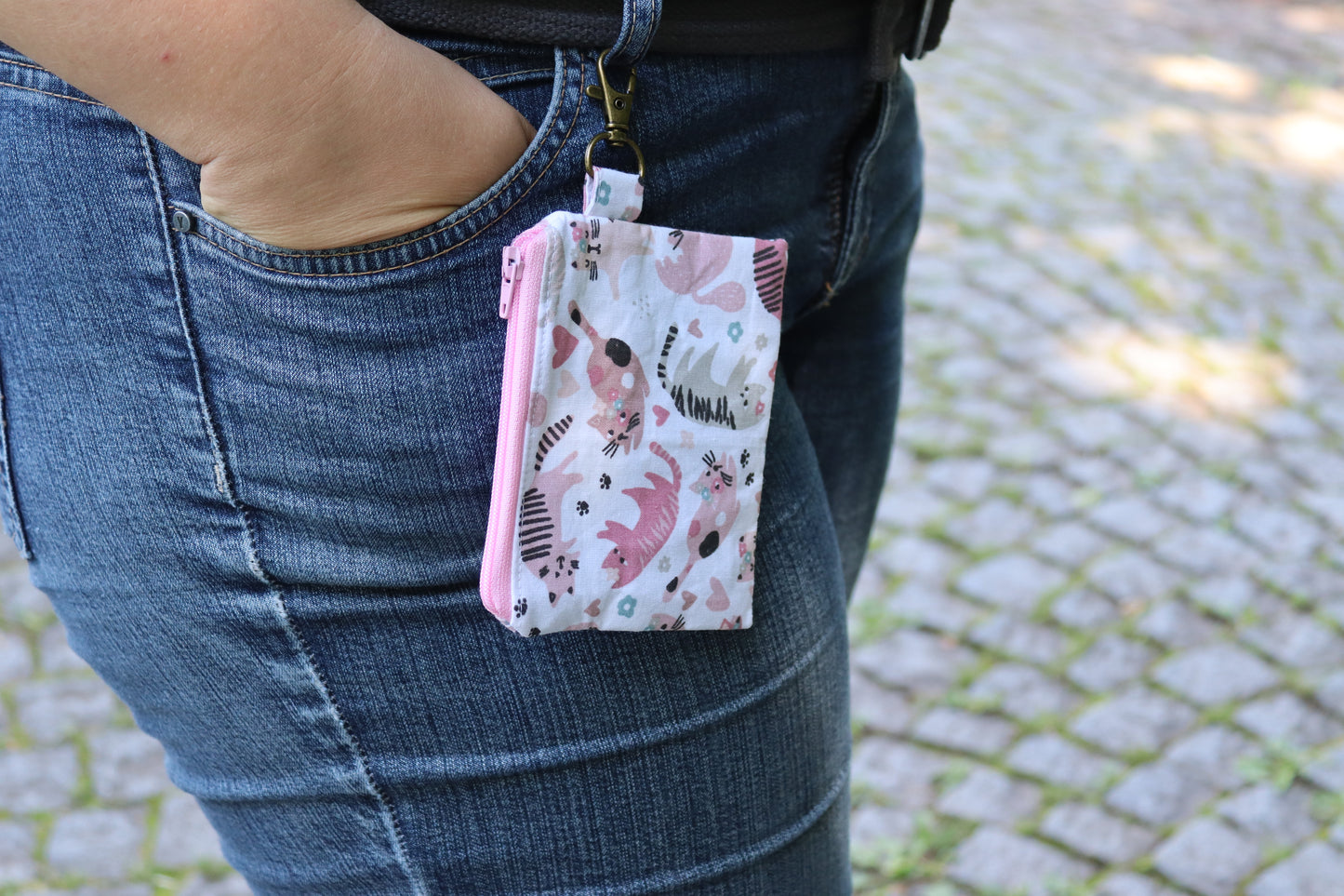 Tragebeispiel Handgenähtes Täschchen (12,5 × 10 cm) mit rosa Katzenmotiven, antikfarbenem Karabiner und rosa Reißverschluss – ideal für Kleinigkeiten unterwegs.
