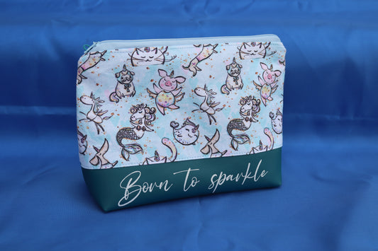 Handgemachte Kosmetiktasche mit hellblauem Stoff und niedlichen Fantasietieren wie Einhörnern, Meerjungfrauen, Mops-Einhörnern und Schweinchen mit Flügeln, darunter petrolgrünes Kunstleder mit der Aufschrift „Born to sparkle“, fotografiert vor blauem Hintergrund.