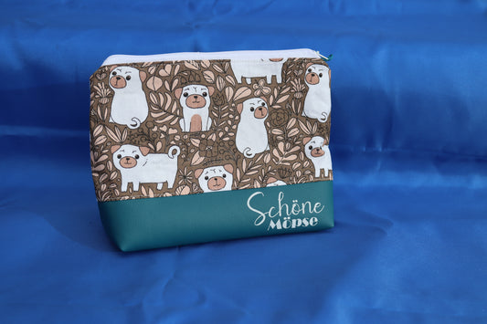 Handgemachte Kosmetiktasche mit braun-weißem Mops-Motiv und der Aufschrift „Schöne Möpse“ auf türkisem Kunstleder, fotografiert vor blauem Hintergrund.