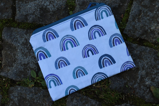 Handgenähtes Täschchen aus weißem Stoff mit unterschidlichen blauen Regenbogenmotiven und dunkelblauem Reißverschluss – kompakt und stilvoll für unterwegs.