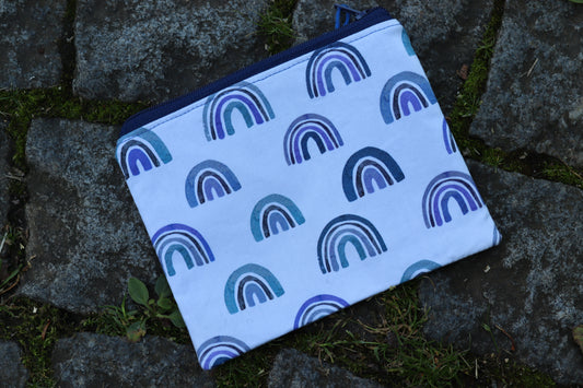 Handgenähtes Täschchen aus weißem Stoff mit unterschidlichen blauen Regenbogenmotiven und dunkelblauem Reißverschluss – kompakt und stilvoll für unterwegs.